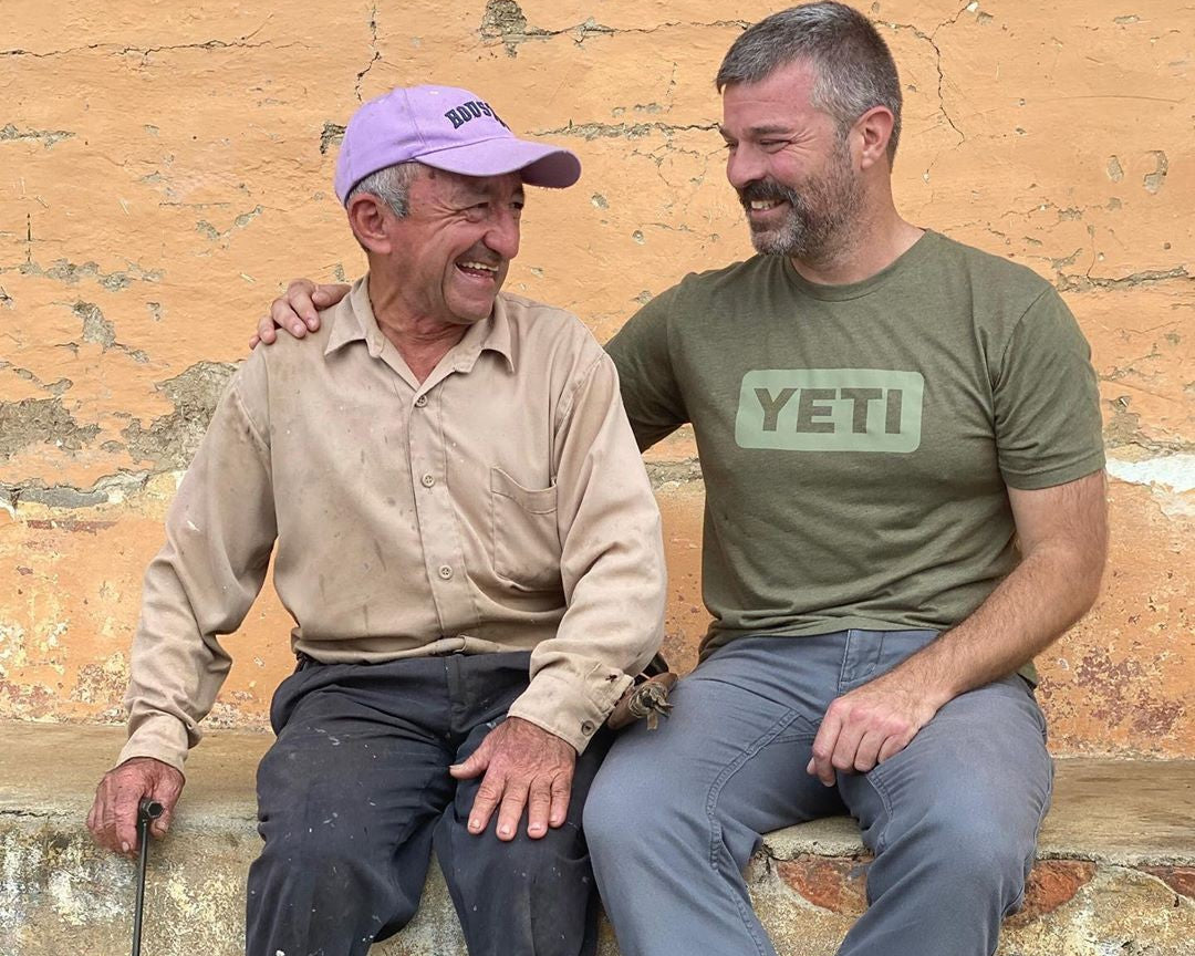 Two men sitting side by side outdoors against a textured orange wall. The man on the right wears a green YETI t-shirt and gray pants, with his arm around the other man, who wears a beige shirt, a purple cap, and black rubber boots.